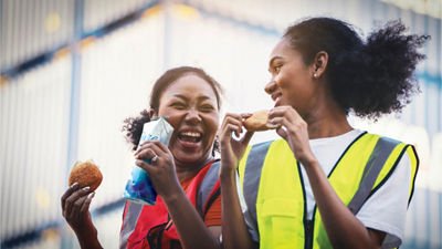 A imagem mostra duas mulheres usando coletes de alta visibilidade, rindo e saboreando um lanche juntas. Uma mulher está segurando uma embalagem cartonada de bebida, enquanto a outra está comendo um sanduíche. Elas estão ao ar livre com um plano de fundo industrial ou de construção visível, sugerindo que estão em um intervalo do trabalho. A atmosfera é alegre e descontraída, destacando um momento de companheirismo.